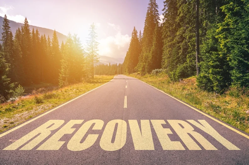 Forest road with the word “Recovery” on the pavement representing the factors that affect the length of rehab and the healing process for addiction recovery.