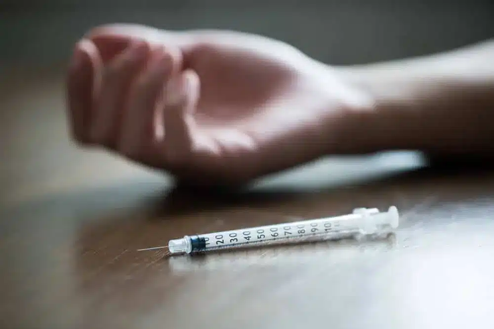 Syringe on table next to an unresponsive person’s hand, representing risks of meth use and possible allergic reaction